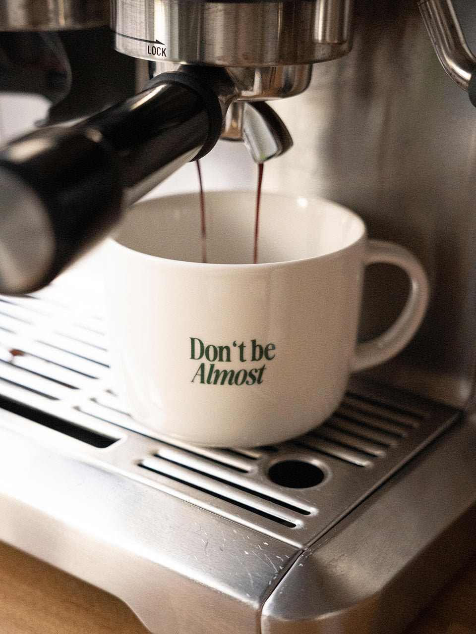Weiße Kaffeetasse mit dem inspirierenden Statement „Don’t be Almost“, aus der frisch gebrühter Espresso aus einer Siebträgermaschine fließt