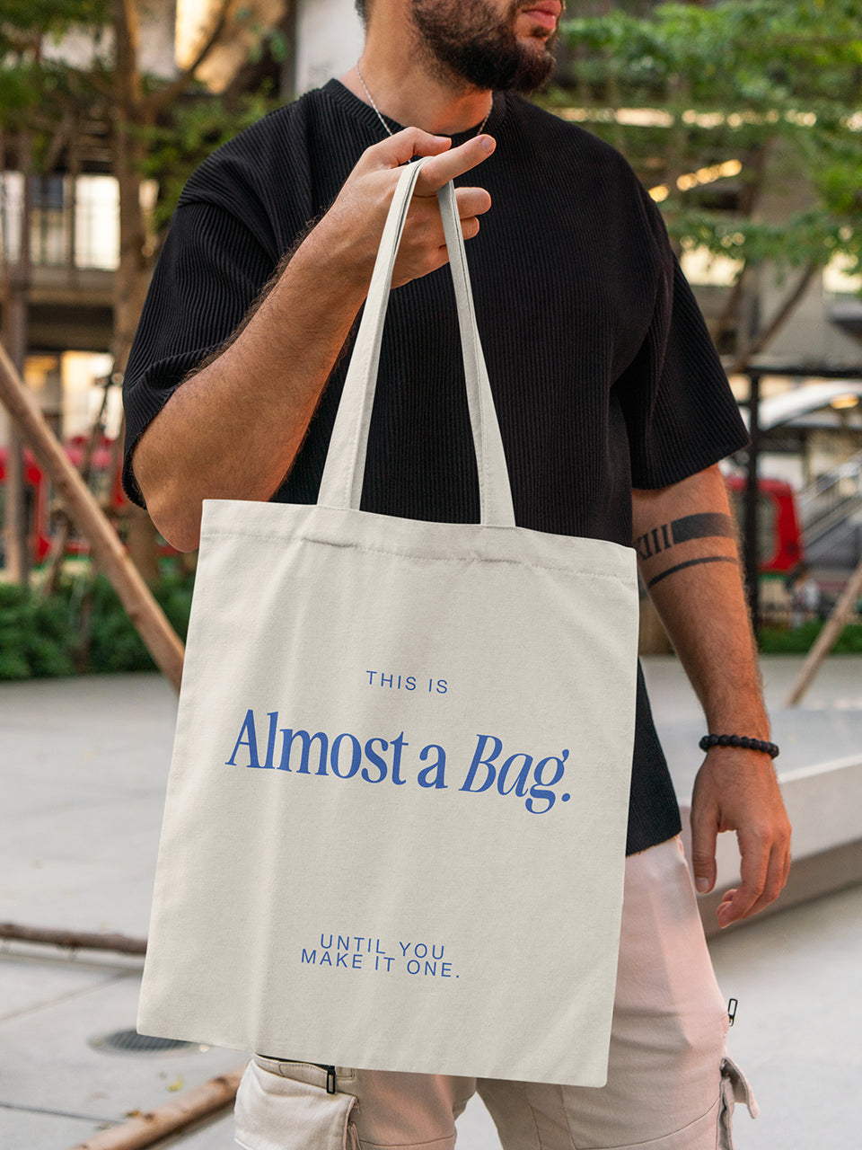 Mann hält eine beigefarbene Totebag mit blauem Textprint „This is Almost a Bag. Until you make it one.“ – ALMOST A Brand