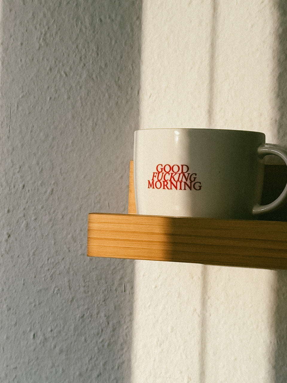 Weiße Tasse mit rotem Aufdruck „GOOD FUCKING MORNING“ von Almost a Brand auf Regalbrett im Sonnenlicht