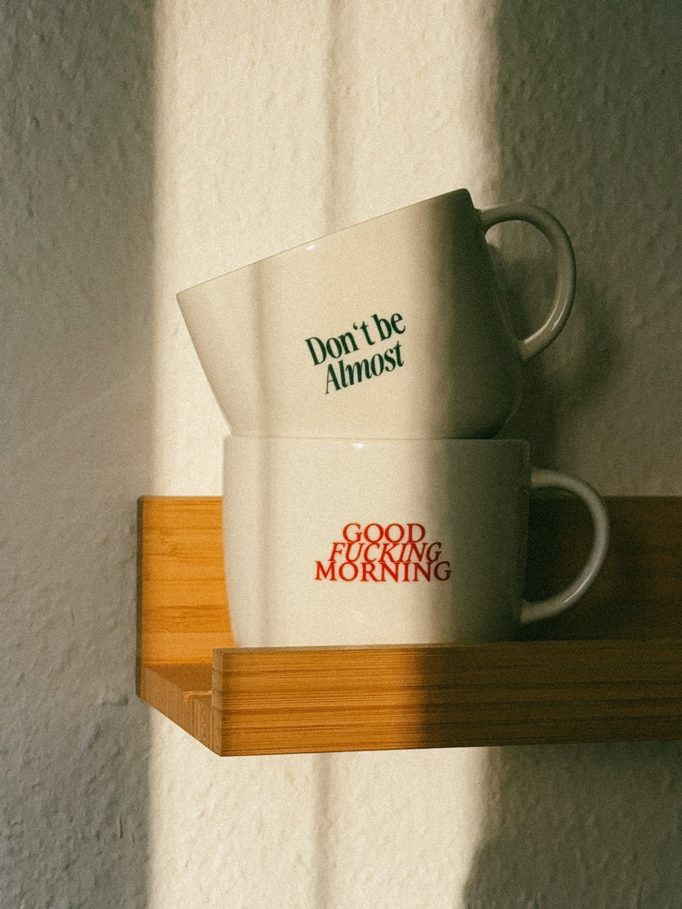Zwei weiße Statement-Tassen von Almost a Brand auf einem Holzregal im warmen Sonnenlicht – mit den Aufdrucken „Don’t be Almost“ und „Good Fucking Morning“