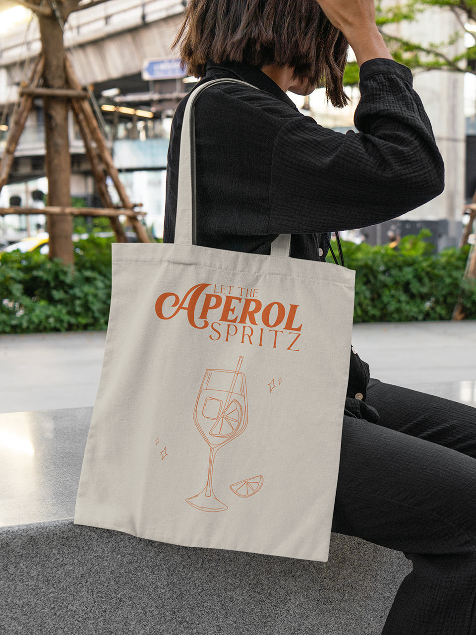 Frau sitzt auf einer Steinbank und trägt eine beigefarbene Stofftasche mit rotem Schriftzug „Let the Aperol Spritz“ und Cocktail-Illustration – ALMOST A BRAND