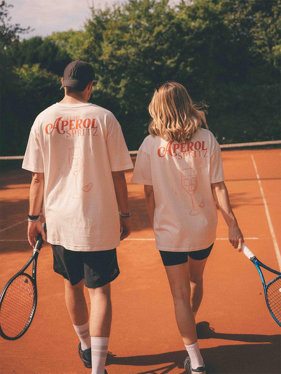 Zwei Personen gehen über einen Tennisplatz, beide tragen weiße Oversized Shirts mit großem „LET THE APEROL SPRITZ“-Print mit einem dezenten Cocktailglas-Motiv