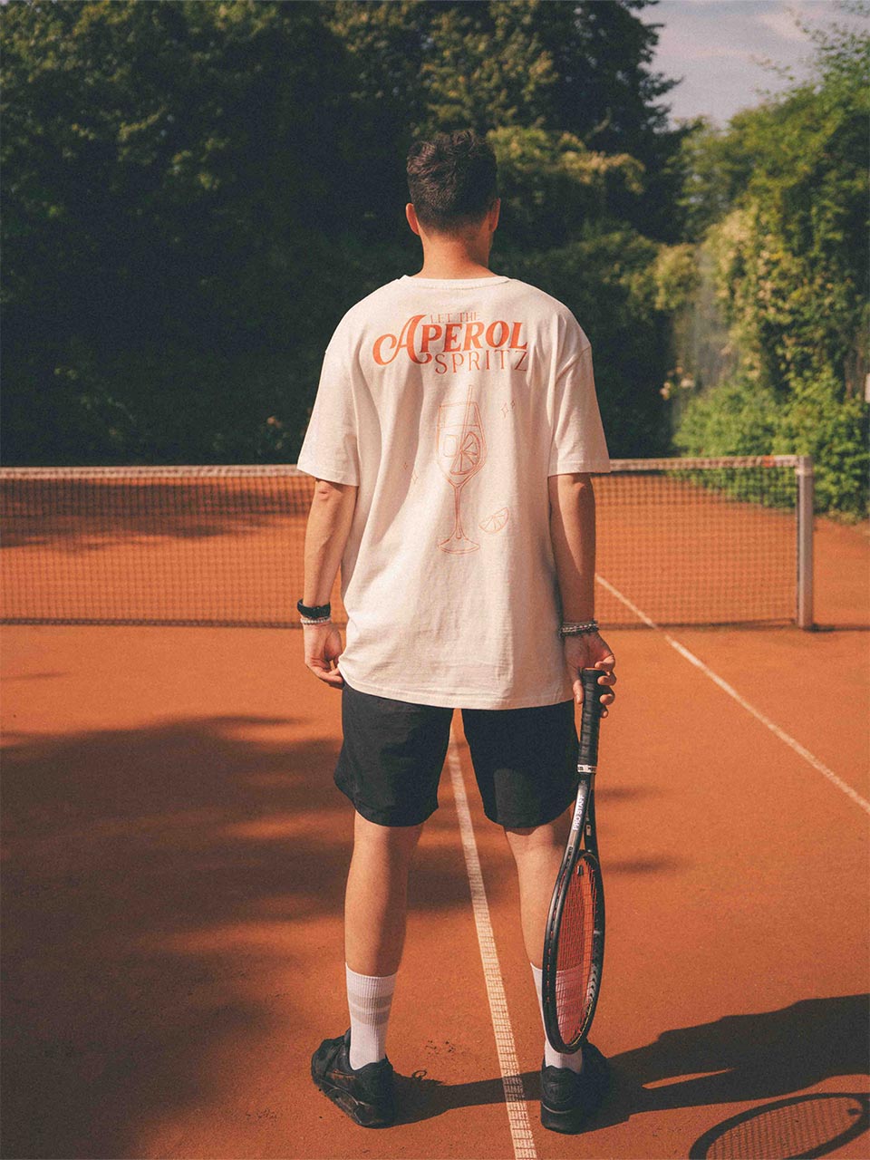 Mann in sportlicher Pose mit Blick auf das Netz; sein T-Shirt zeigt auf dem Rücken einen orangefarbenen Schriftzug „LET THE APEROL SPRITZ“ mit einem Cocktailglas-Motiv gefüllt mit Eiswürfel und Orangenscheibe.