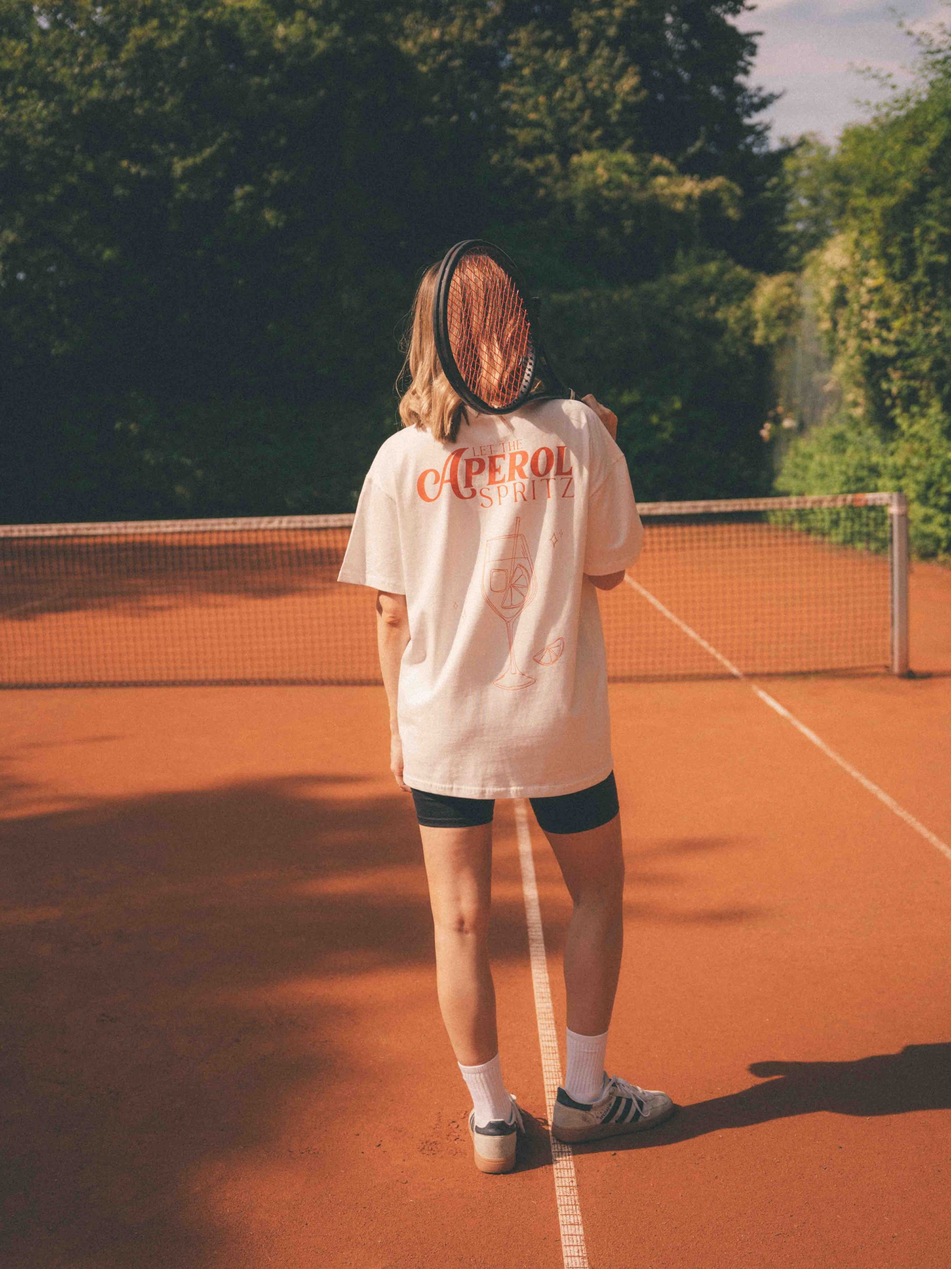 Frau auf dem Tennisplatz mit dem Rücken zur Kamera; ihr weißes T-Shirt trägt ein dezentes Cocktailglas-Motiv mit Eiswürfeln und einer Orangenscheibe mit dem Text „LET THE APEROL SPRITZ“.