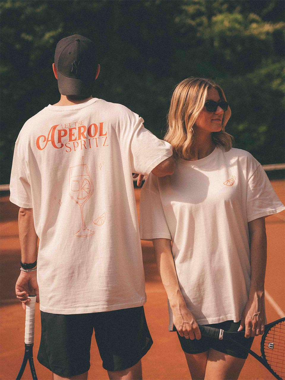 Ein Mann und eine Frau auf dem Tennisplatz; das T-Shirt des Mannes zeigt einen Cocktail mit Eiswürfel und orangenscheibe mit dem Spruch „LET THE APEROL SPRITZ“, die Frau trägt das gleiche Shirt nach vorne gerichtet, mit einer kleinen Orangenscheibe auf der linken Brust, locker über Shorts.