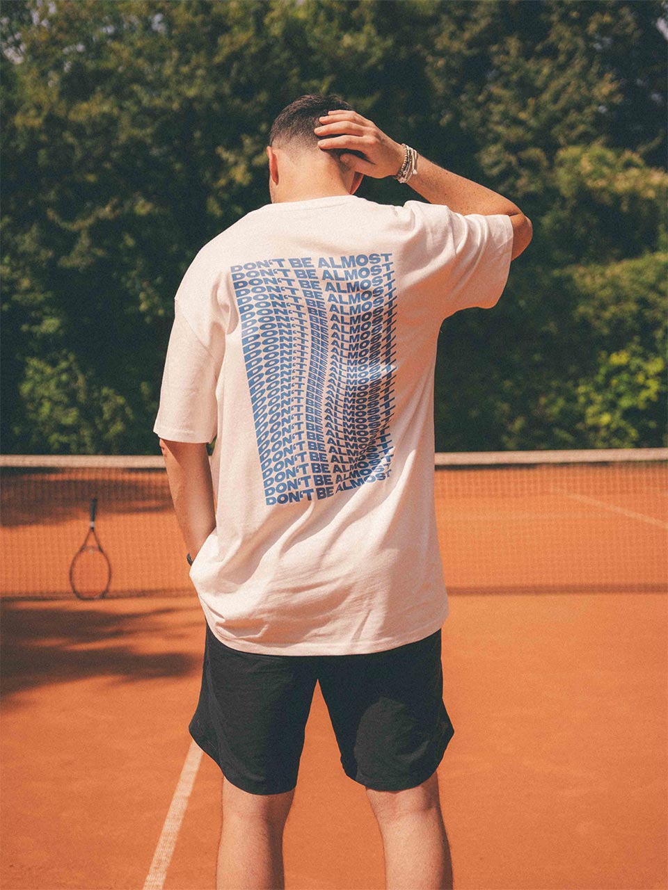 Mann steht auf einem Tennisplatz und hat die rechte Hand am Hinterkopf. Auf seinem weißen T-Shirt ist rückseitig in Blau mehrfach der Schriftzug „Don’t Be Almost“ im verzerrten Stil zu sehen.