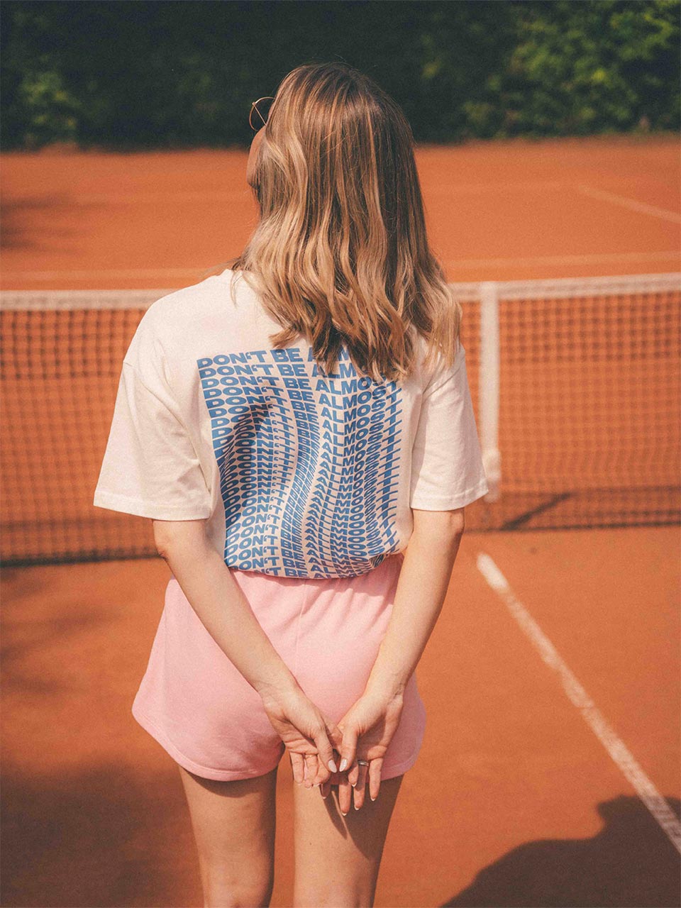 Rückansicht einer Frau mit welligem, blonden Haar auf einem Tennisplatz. Sie trägt ein weißes T-Shirt mit einem großflächigen blauen Schriftzug „Don’t Be Almost“ auf dem Rücken und eine rosafarbene Shorts.