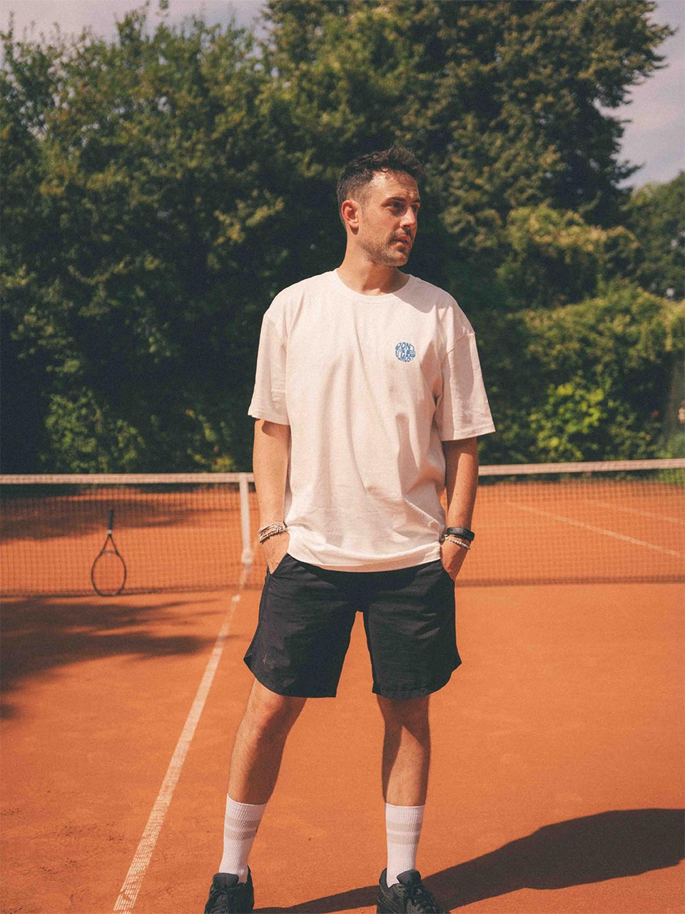 Mann steht entspannt mit Händen in den Hosentaschen auf einem Sandtennisplatz. Er trägt ein weißes T-Shirt mit kleinem blauen „Don’t Be Almost“-Print auf der Brust, dazu schwarze Shorts und Tennissocken.