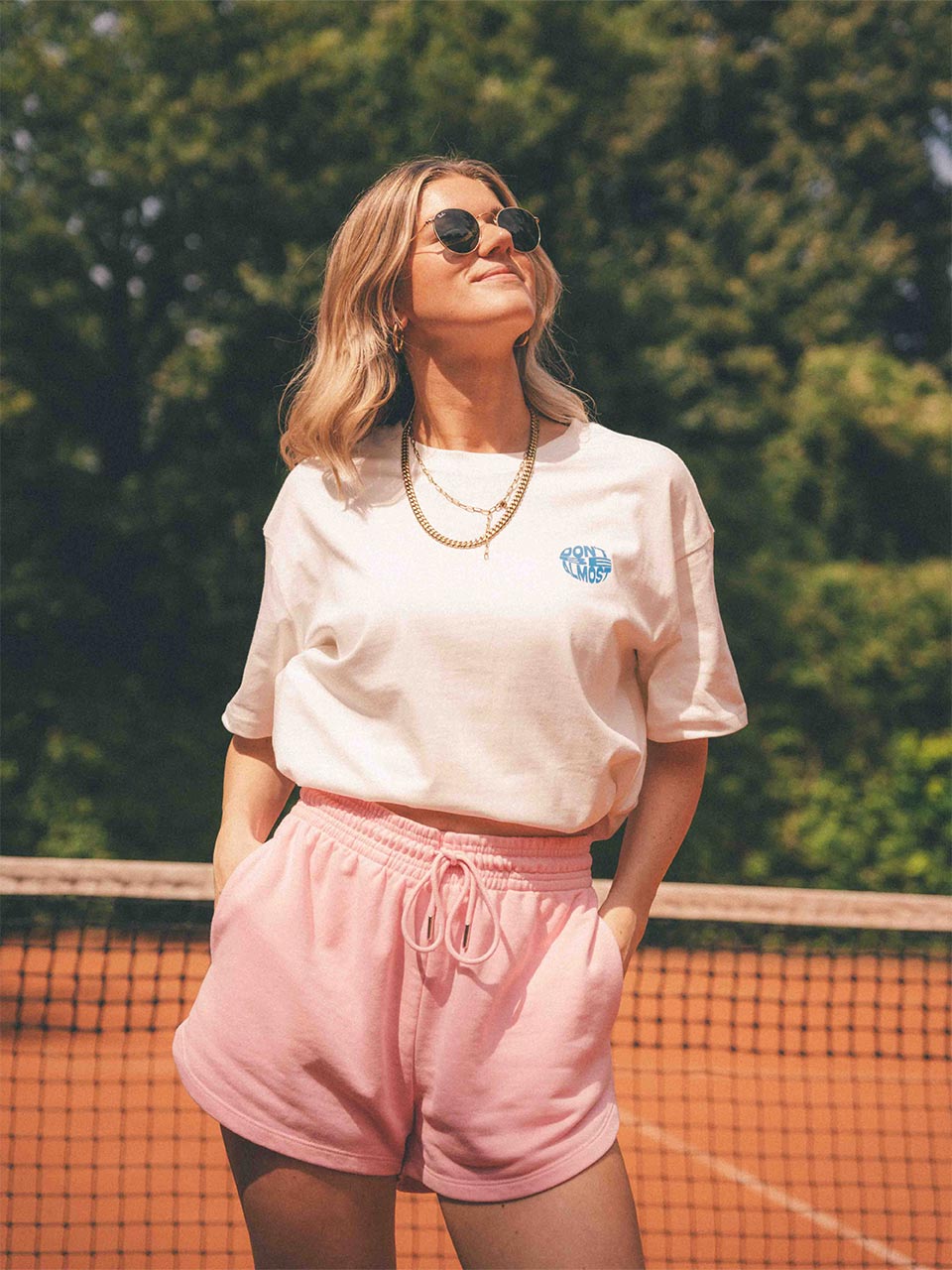 Frau mit blonden Haaren steht auf einem Tennisplatz, trägt ein weißes T-Shirt mit kleinem blauen Aufdruck „Don’t Be Almost“ auf der Brust und eine rosafarbene Shorts, schaut in die Sonne und lächelt leicht.
