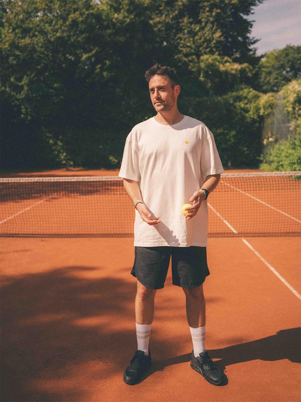 Mann steht auf einem Tennisplatz und trägt ein weißes Oversized-T-Shirt mit kleinem gelbem Smiley-Ei-Print auf der Brust - dazu dunkle kurze Sporthose