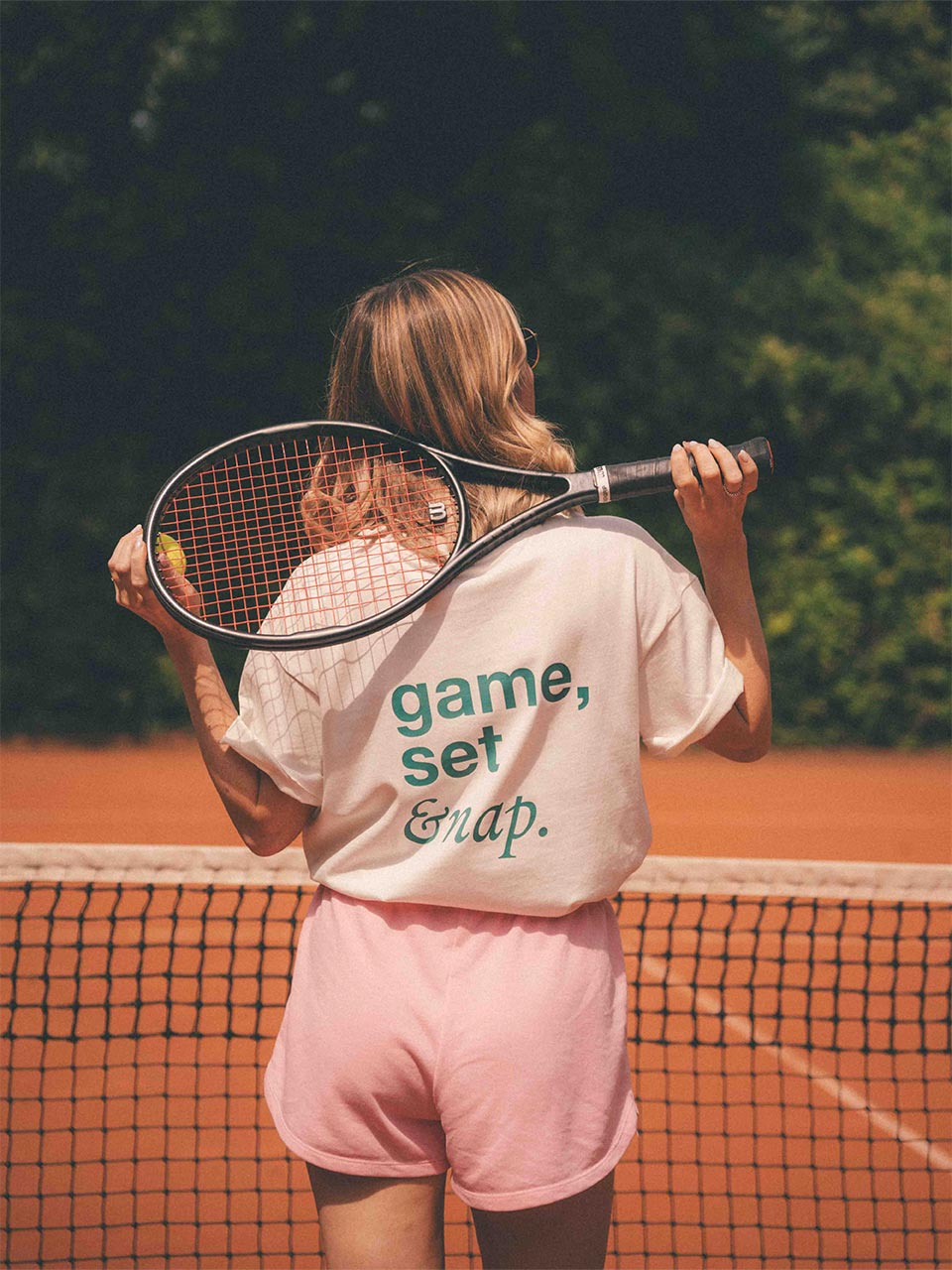 Frau auf dem Tennisplatz mit einem Tennisschläger über der Schulter, trägt ein weißes Almost a Brand T-Shirt mit grünem Rückendruck „game, set & nap.“ – kombiniert mit rosa Shorts und Sonnenbrille.