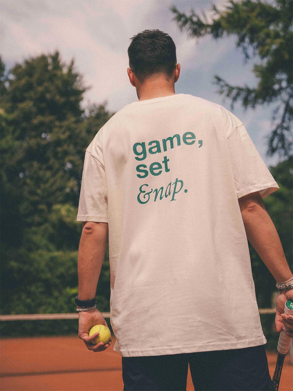 Mann auf dem Tennisplatz trägt ein weißes Almost a Brand T-Shirt mit großem Rückendruck „game, set & nap.“ in Grün. dazu hält er einen tennisschläger und einen tennisball in seinen Händen.