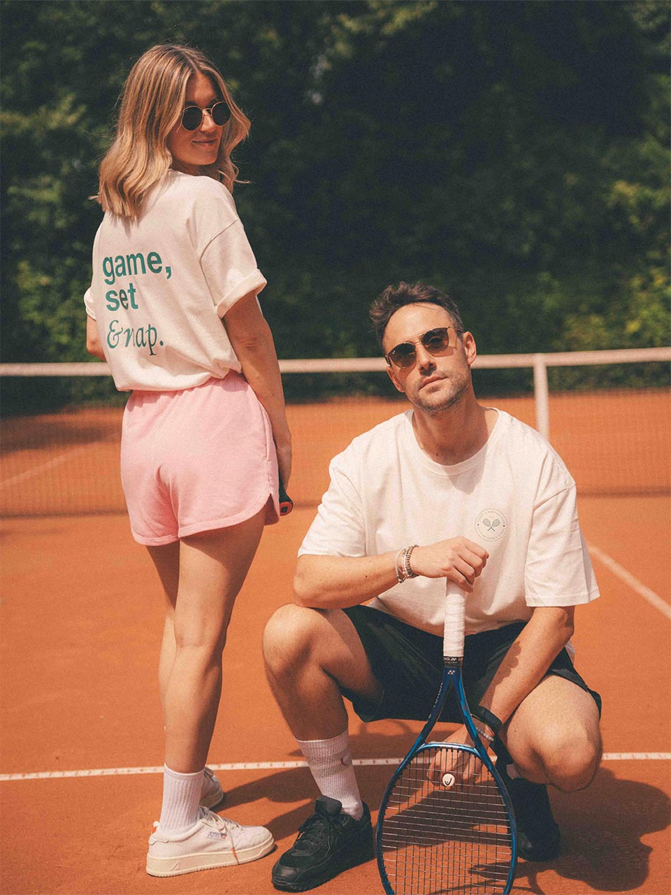 Zwei Personen in weißem Almost a Brand T-Shirts mit Tennis-Motiven posieren auf dem Tennisplatz: Frau mit Rückendruck „game, set & nap.“, Mann mit kleinem Tennis-Logo auf der Brust