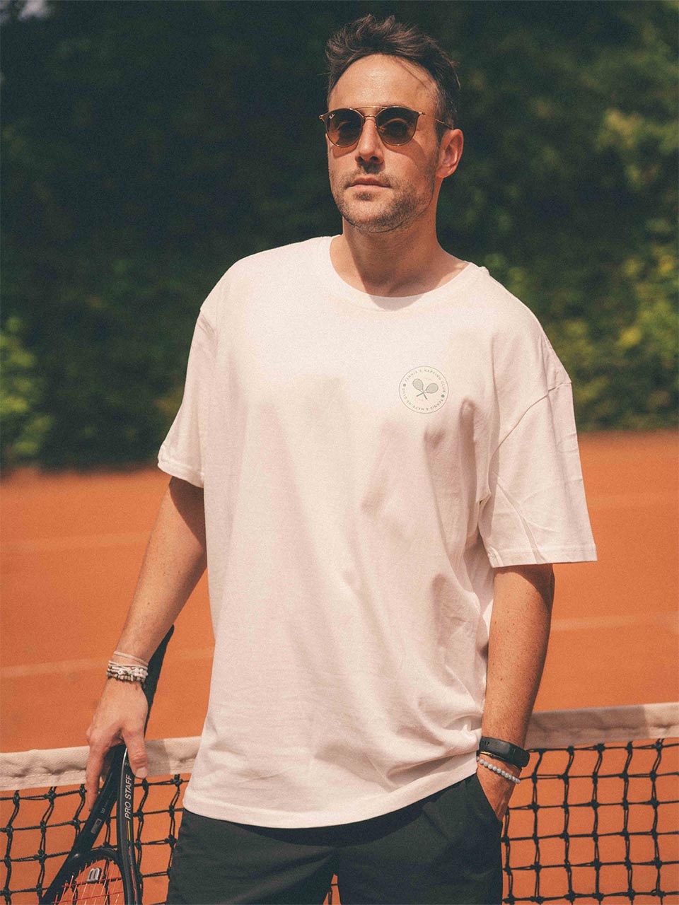 Männliches Model auf dem Tennisplatz trägt das „game, set & nap.“ T-Shirt von Almost a Brand in Weiß mit kleinem Tennis-Logo auf der Brust – zusammen mit dunklen Shorts, Tennischläger, Armbändern und Sonnenbrille.