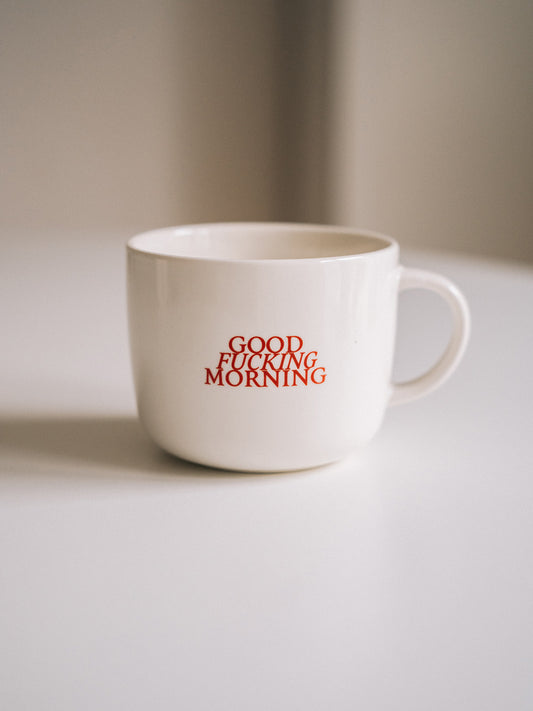 Weiße Kaffeetasse mit rotem Statement-Aufdruck „GOOD FUCKING MORNING“, fokussiert fotografiert auf hellem Untergrund.