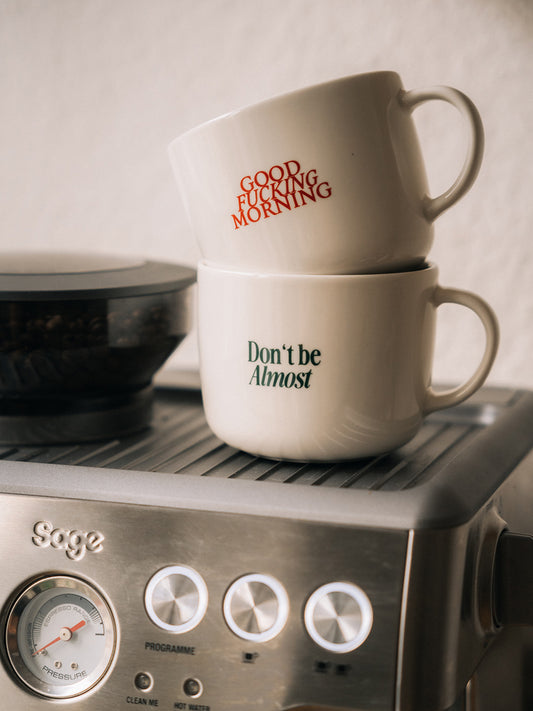 Zwei weiße Statement-Kaffeetassen von ALMOST A BRAND mit den Aufdrucken „GOOD FUCKING MORNING“ und „Don’t be Almost“, gestapelt auf einer Siebträgermaschine