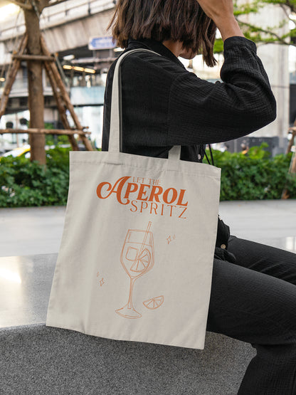 Frau sitzt auf einer Steinbank und trägt eine beigefarbene Stofftasche mit rotem Schriftzug „Let the Aperol Spritz“ und Cocktail-Illustration – ALMOST A BRAND
