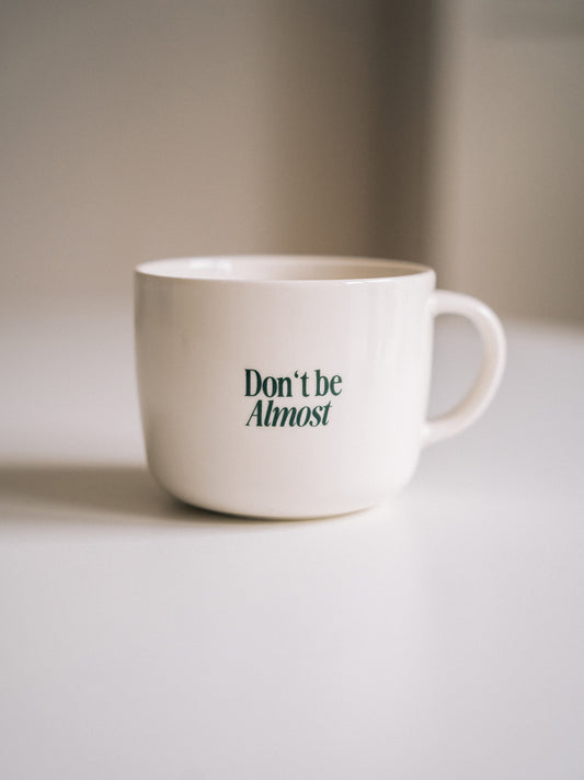 Weiße "Don´t be Almost"-Kaffeetasse mit grünem Statement-Aufdruck „Don’t be Almost“, fokussiert fotografiert auf hellem Untergrund.