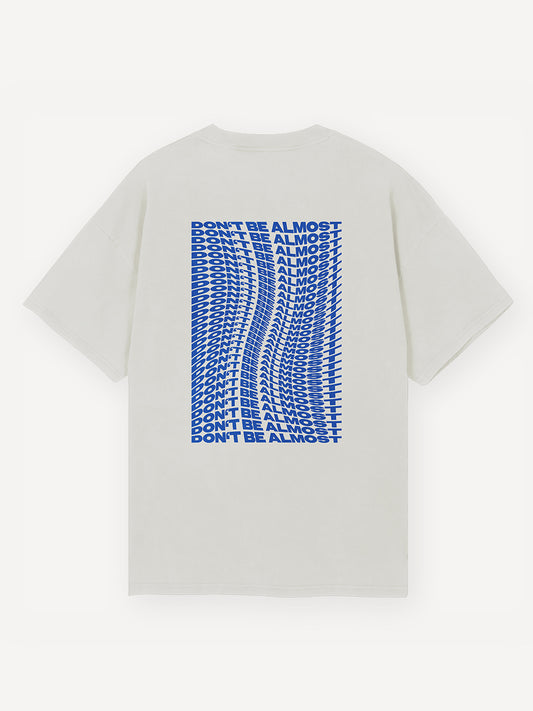 Weiße Rückseite eines T-Shirts mit einem leuchtend blauen, verzerrten Schriftzug in mehrfacher Wiederholung: „DON’T BE ALMOST“, zentriert aufgedruckt in blockartiger Typografie.