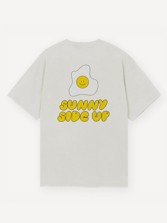Großflächige Darstellung eines lächelnden Spiegeleis, darunter befindet sich ein auffälliger gelber Schriftzug in verspieltermSchrift: „SUNNY SIDE UP“ auf einem weißen Oversized T-Shirt