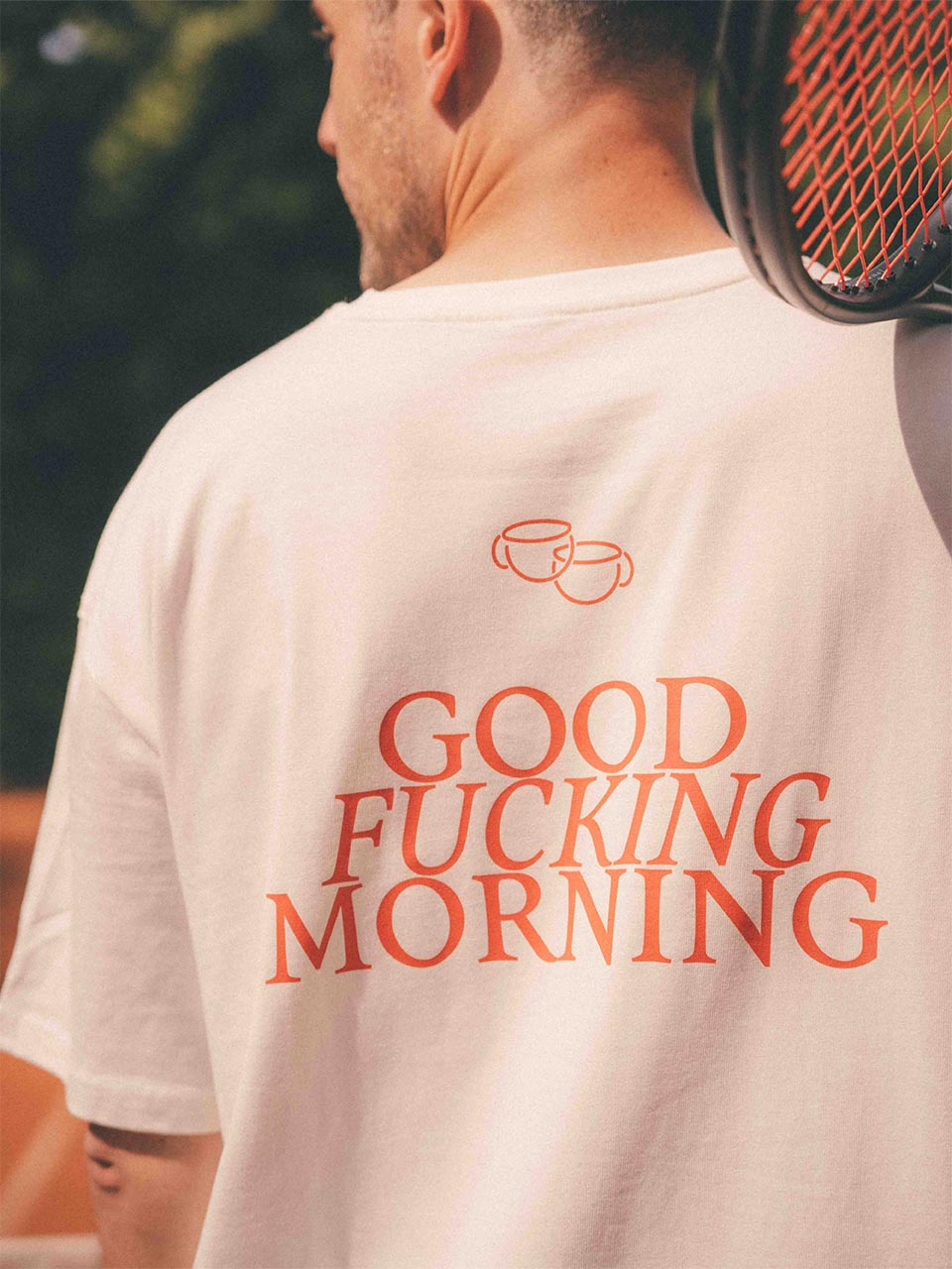 Rückenansicht eines Mannes im weißen T-Shirt mit großem roten Print: „GOOD FUCKING MORNING“, darüber ein ikonischer Linien-Print zweier Kaffeetassen. Ein Tennisschläger liegt lässig auf seiner Schulter, im Hintergrund ist verschwommenes Grün sichtbar.