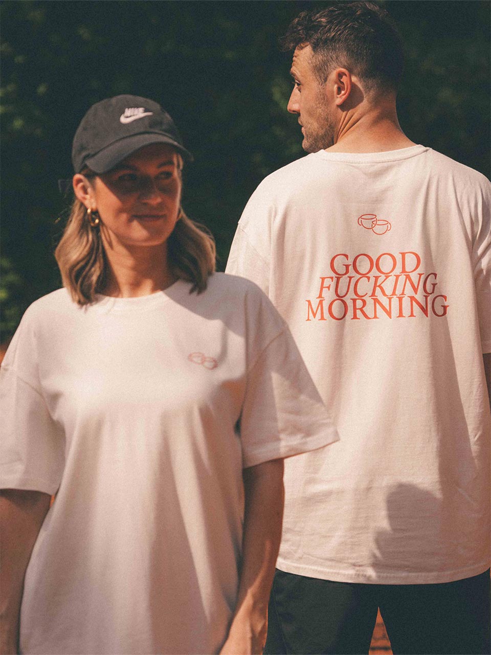 Frau und Mann stehen im Sonnenlicht nebeneinander. Beide tragen ein weißes Oversize-T-Shirt mit minimalistischem roten Brust-Print aus zwei gezeichneten Kaffeetassen. Die Frau blickt zur Seite, der Mann steht leicht gedreht mit dem Rücken zur Kamera und zeigt den großen Rückendruck mit dem Schriftzug „GOOD FUCKING MORNING“ in roter Schrift.