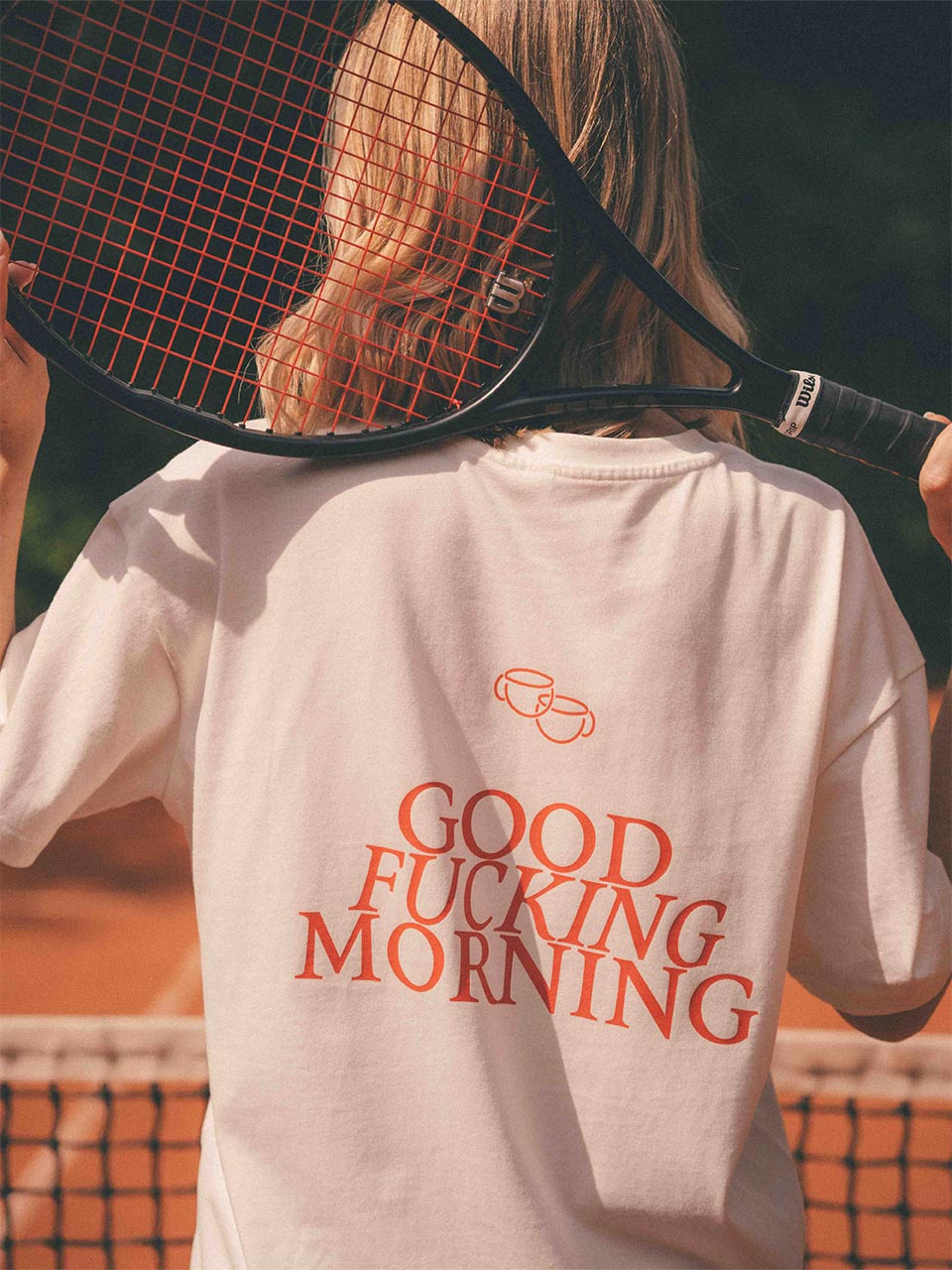 Frau auf dem Tennisplatz mit Tennisschläger über den Schultern trägt ein weißes Oversized-T-Shirt mit großem roten Rückenprint „GOOD FUCKING MORNING“ und zwei Kaffeetassen darüber 