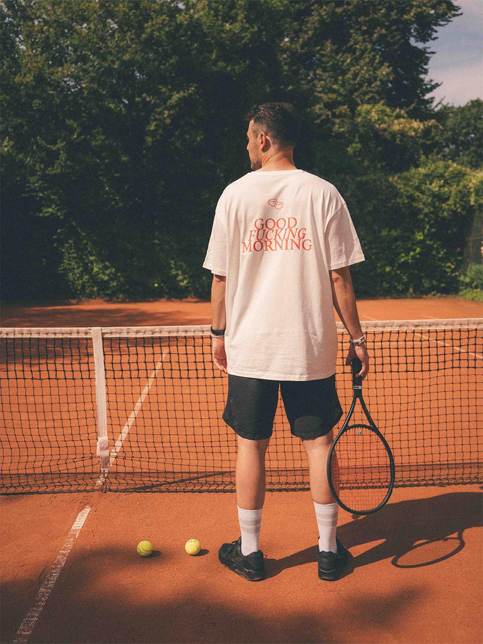 Rückansicht eines Mannes mit Tennisschläger in der Hand auf einem Tennisplatz im weißen Oversized-T-Shirt von Almost a Brand mit auffälligem roten Statement-Print „GOOD FUCKING MORNING“ und kleinen Kaffeetassen darüber