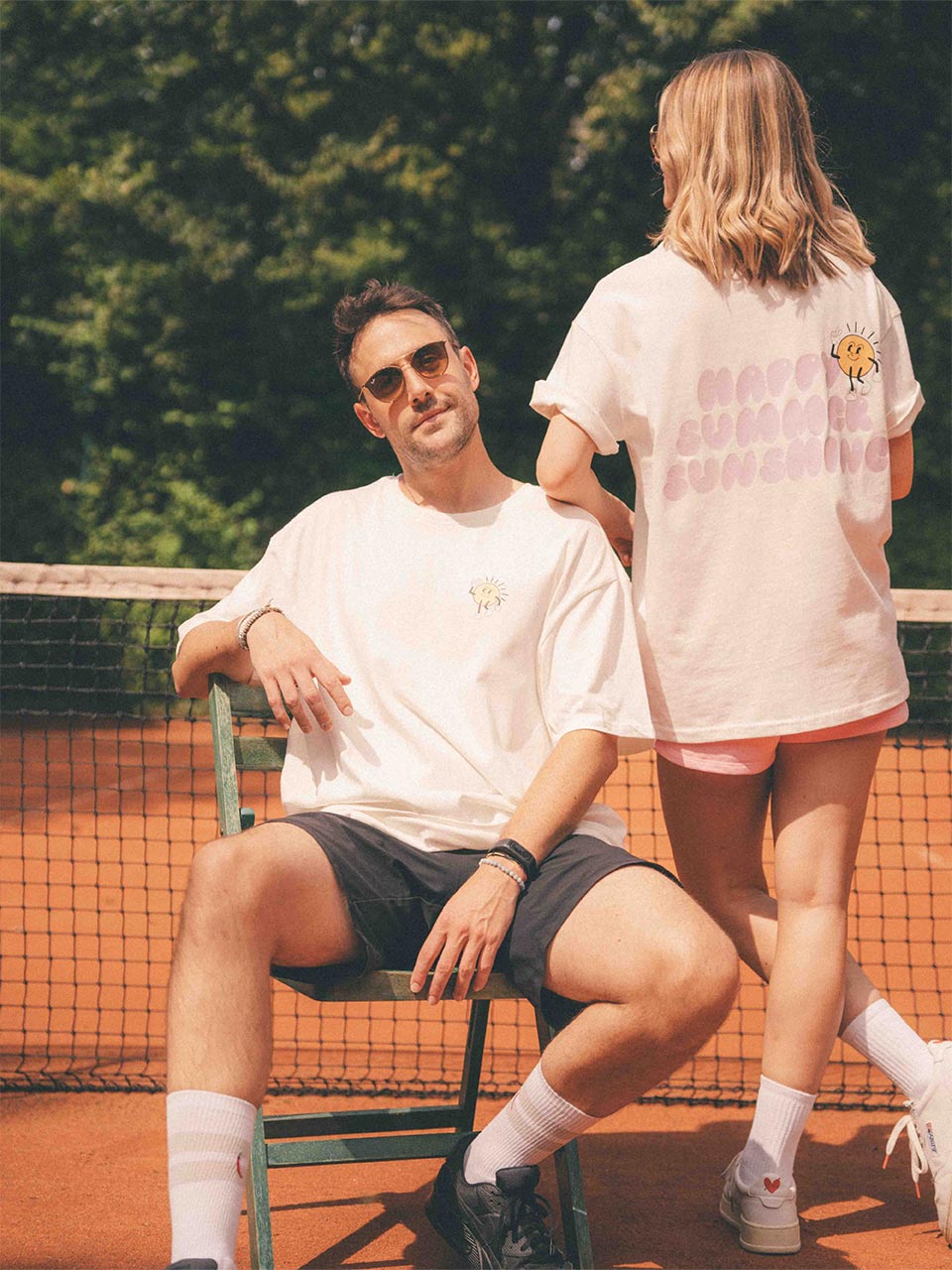 Mann und Frau tragen Almost a Brand T-Shirts aus der „Happy Summer Sunshine“-Kollektion auf einem Tennisplatz – er mit kleinem Sonnen-Print auf der Brust, sie mit großem Rückendruck „HAPPY SUMMER SUNSHINE“ und Cartoon-Sonne