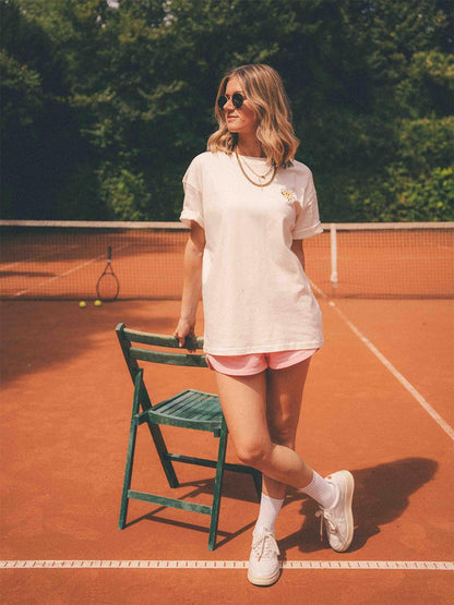 Frau steht auf einem Tennisplatz in einem weißen T-Shirt mit kleiner Sonnen-Grafik auf der Brust, kombiniert mit rosa Shorts, weißen Sneakers und runder Sonnenbrille 