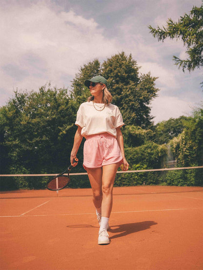 Weibliches Model auf dem Tennisplatz trägt das „game, set & nap.“ T-Shirt von Almost a Brand in Weiß mit kleinem Tennis-Logo auf der Brust – kombiniert mit rosa Shorts, grüner Cap, goldenen Ketten und Sonnenbrille. 