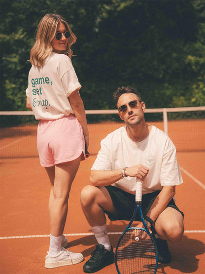 Zwei Personen in weißem Almost a Brand T-Shirts mit Tennis-Motiven posieren auf dem Tennisplatz: Frau mit Rückendruck „game, set & nap.“, Mann mit kleinem Tennis-Logo auf der Brust